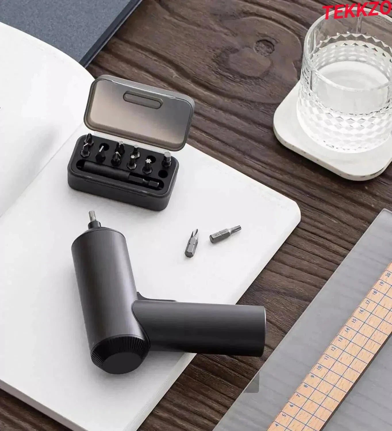 Compact black cordless electric screwdriver with multiple bits on a wooden desk with glass of water